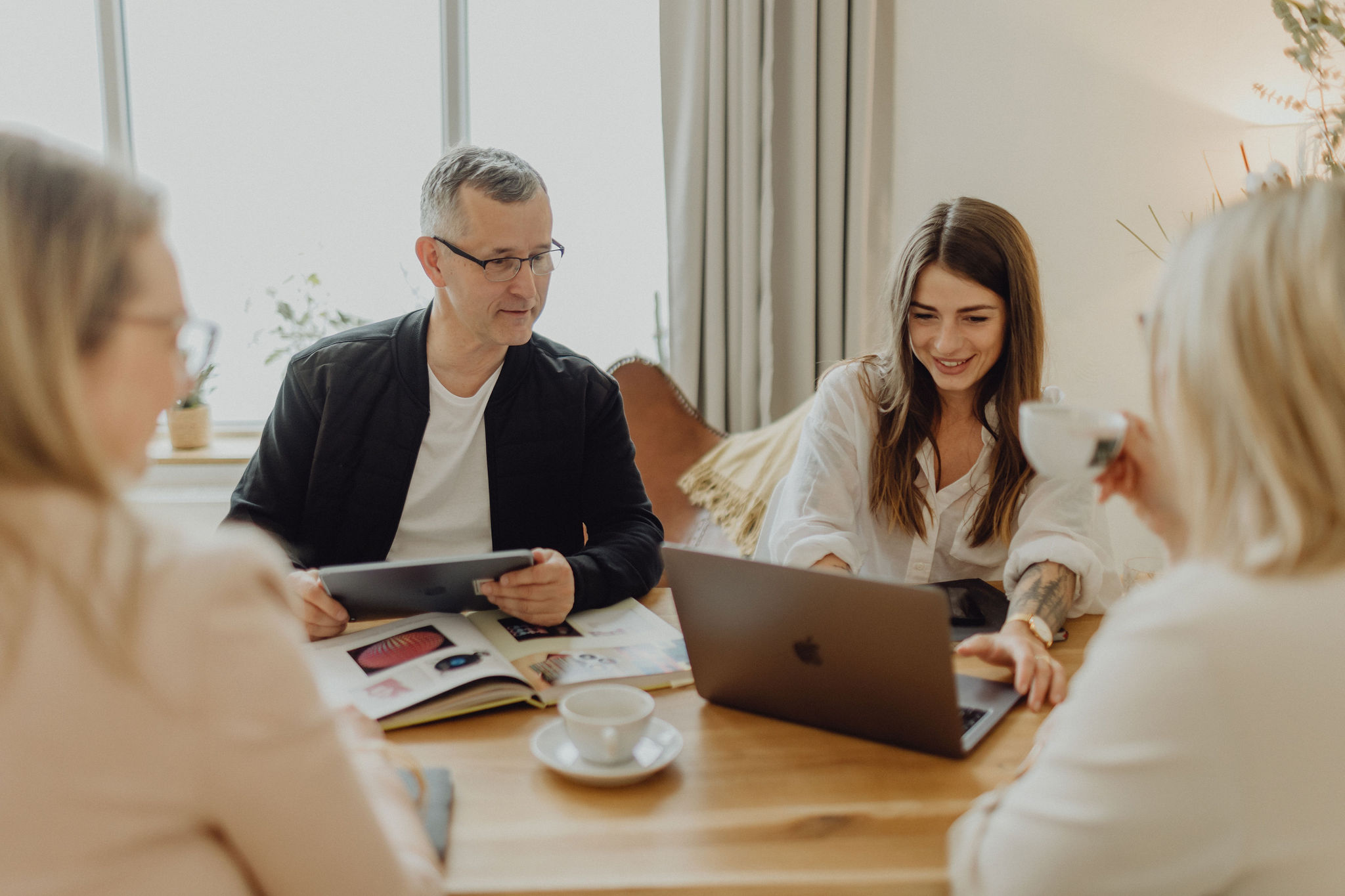 Teammeeting an einem Holztisch, vier Personen arbeiten mit Laptop, Tablet und B&uuml;chern in einem modernen und entspannten B&uuml;ro.