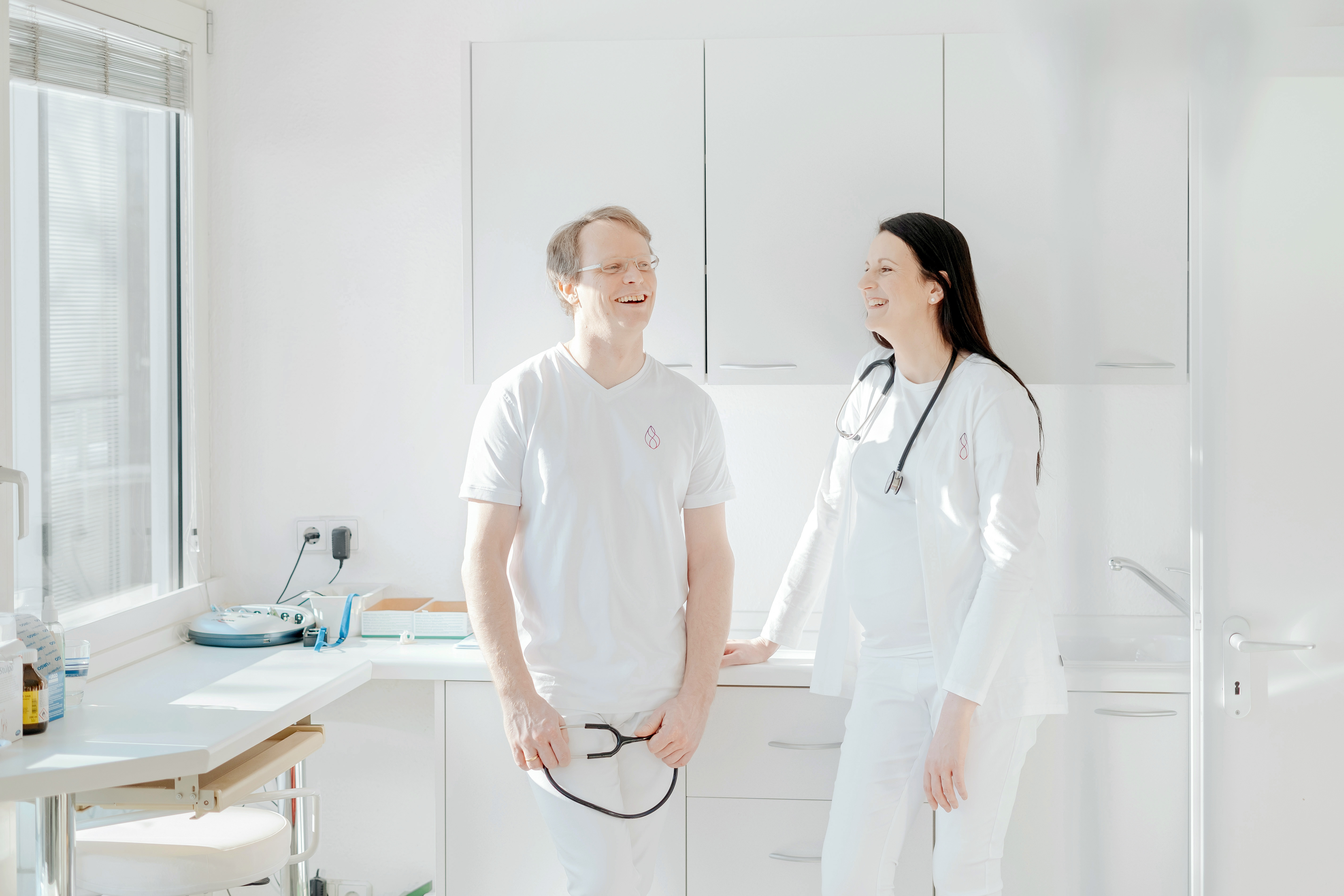 Zwei &Auml;rzte in wei&szlig;er Kleidung l&auml;chelnd in einer hellen Arztpraxis, modernes und freundliches Gesundheitsumfeld.