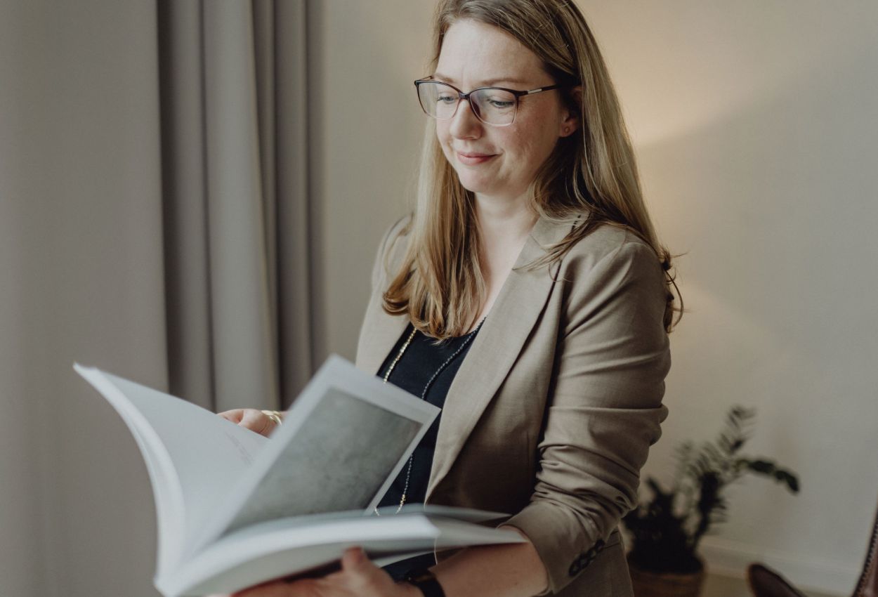 Geschäftsfrau mit Brille und Blazer liest ein Buch, professionelles Porträt in einem stilvollen, modernen Büroambiente.