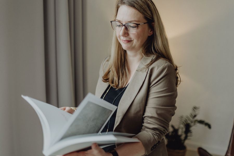 Geschäftsfrau mit Brille und Blazer liest ein Buch, professionelles Porträt in einem stilvollen, modernen Büroambiente.