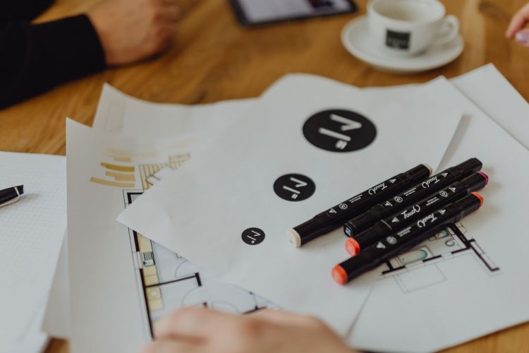 Designskizzen und Marker auf einem Holztisch, mit Kaffeetasse im Hintergrund, Fokus auf kreatives Arbeiten und Planung.