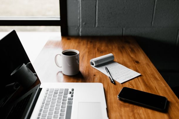 Minimalistischer Arbeitsplatz mit Laptop, Notizblock, Stift, Smartphone und Kaffeetasse auf Holztisch in natürlichem Licht.